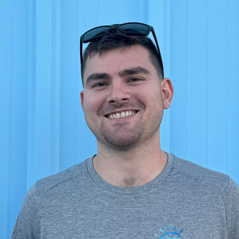 Team member smiling with sunglasses on head in front of blue wall.